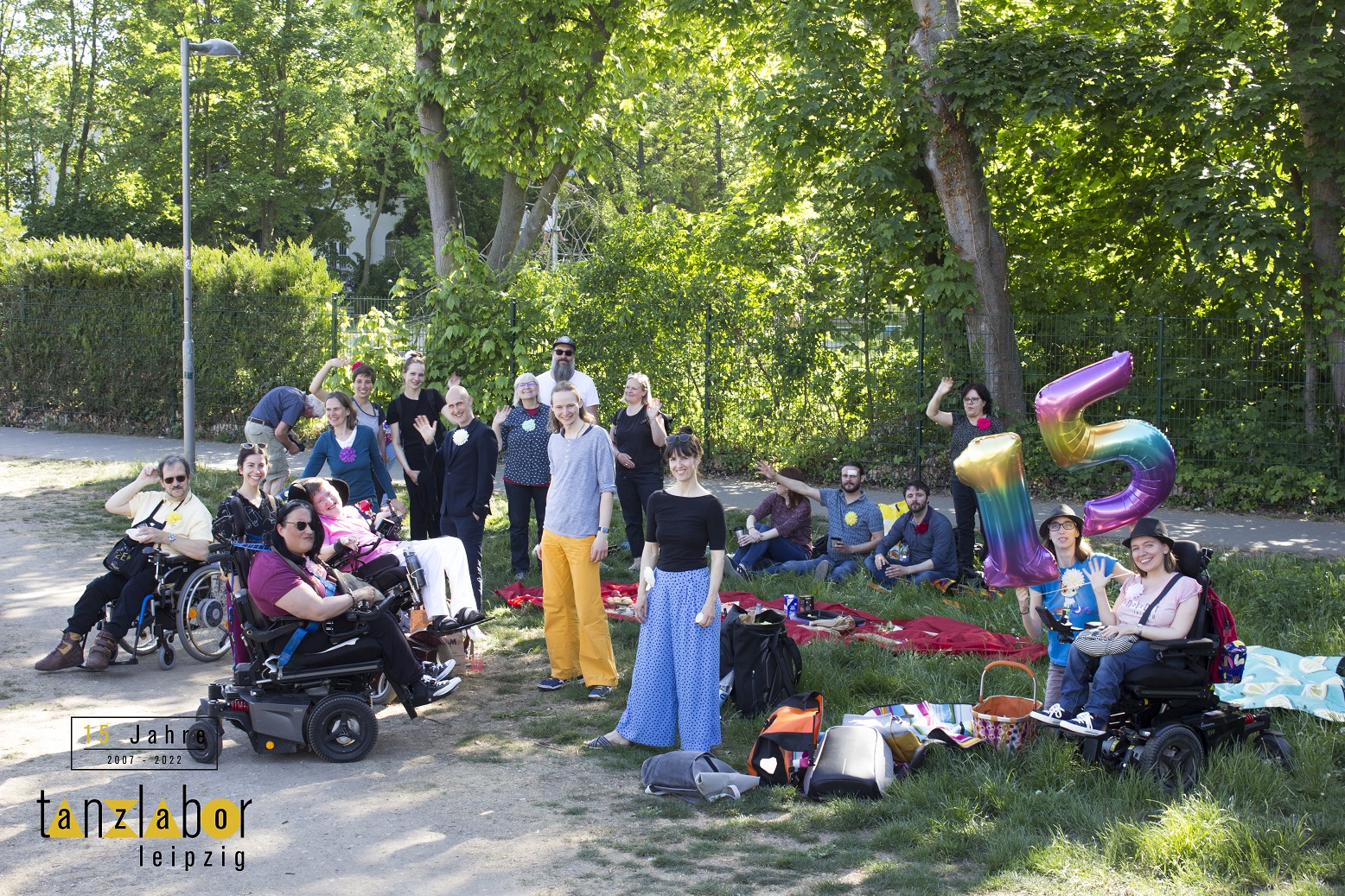 Freunde und Mitwirkende vom Tanzlabor winken in die Kamera. Ein Picknick auf einer Wiese ist zu sehen. Zwei Ballons zeigen die Zahl 15.