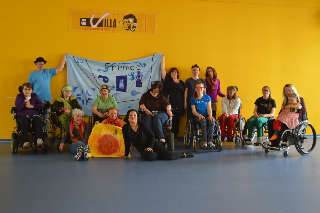 Foto vom Freien Tanzen. Alle Teilnehmenden stehen vor einer gelben Wand und schauen in die Kamera. Einige Personen sitzen im Rollstuhl. Zwei Personen halten ein Banner hoch mit Zeichnungen und dem Wort "Fremde"